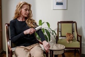 A woman cleaning her clothes with a lint roller while a ginger cat sits nearby, indoors.